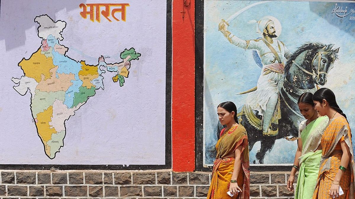 Indian women walk past a political map of the country and an image of Maratha warrior king Chatrapati Shivaji painted on a school building wall